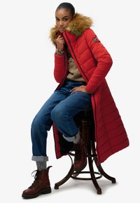 Rød puffet jakke med pelsforede hætte, parret med en tan sweater, blå jeans og brune snørebåndsstøvler, siddende på en træstol.