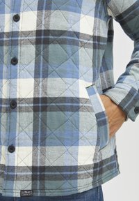 Kockás, steppelt flanel ing kék, zöld és fehér színekben. Fekete gombokkal, két oldalsó zsebbel és címkével az alsó szegélyen.