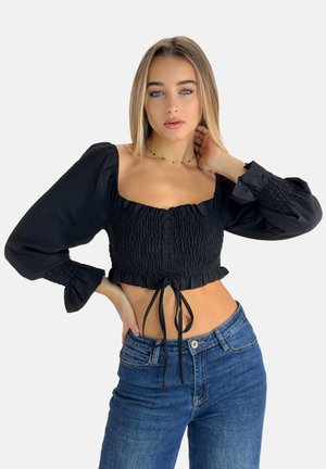 Schwarze, kurze Bluse mit gerüschter Gestaltung, carréförmigem Ausschnitt und Puffärmeln. Kombiniert mit hochtaillierten blauen Jeans. Glatte Textur.