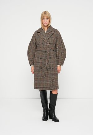 Ruskean ja kermanvärisen ruudullinen trench-takki, jossa on kaksirivinen design, leveät hihat ja vyötäröllä oleva vyö. Mustat polvisukat saappaat täydentävät asua.