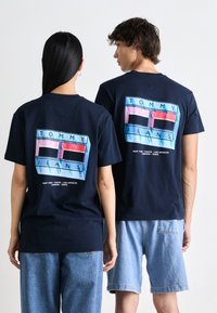 Mørkeblå bomulds t-shirts med en grafisk design, der viser "TOMMY JEANS" i pink, blå og sort. Model iført lyse denimshorts.