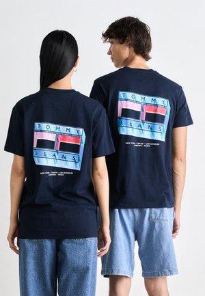 T-shirts en coton bleu marine avec un design graphique affichant "TOMMY JEANS" en rose, bleu et noir. Modèle portant un short en denim clair.