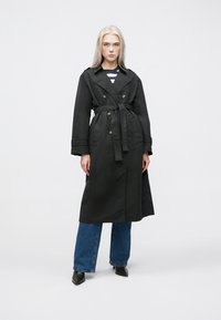 Črni trench plašč z dvosmernimi gumbi, pasom, širokimi ovratniki in stranskimi žepi, kombiniran z modrimi kavbojkami in črnimi koničastimi čevlji.