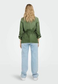 Camicia verde leggera con vita regolabile, maniche lunghe arrotolate, abbinata a jeans larghi blu chiaro e sneakers bianche.