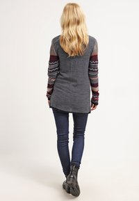 Szary sweter z wzorzystymi rękawami w kolorze bordowym, granatowym i kremowym. Dzianinowy materiał; dłuższy z tyłu z bocznymi rozcięciami. Noszony z ciemnymi dżinsami i czarnymi botkami.