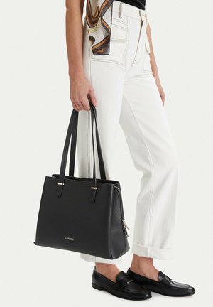 Osoba ve bílých kalhotách a černých mokasínách drží černou kabelku Lasocki s dvěma úchyty a viditelným detailem zipu.