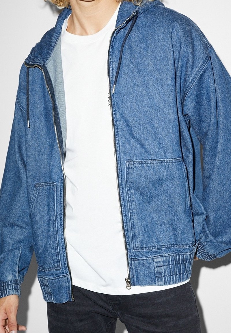 Blå denimjacka med huva, framdragkedja och elastiska ärmmuddar. Har två stora fickor fram och en avslappnad passform.