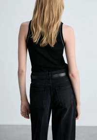 Top dintr-o țesătură ribbed neagră, asociat cu blugi negri din denim cu talie înaltă, având o curea din piele netedă. Părul lung și drept este vizibil.