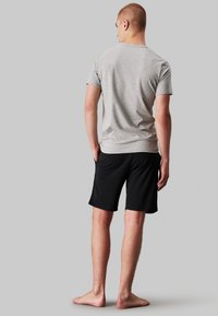 Gray short-sleeve t-shirt made of cotton, paired with black stretch shorts. The model has bare feet, facing away against a gray background.