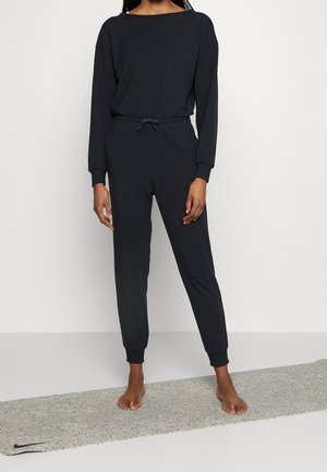 Femme debout pieds nus sur un tapis de yoga gris, portant une combinaison noire à manches longues, avec des chevilles resserrées et une taille à cordon de serrage.