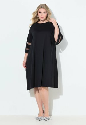 Model wearing a black dress with sheer sleeves, standing against a white background. She accessorizes with silver high-heeled shoes.