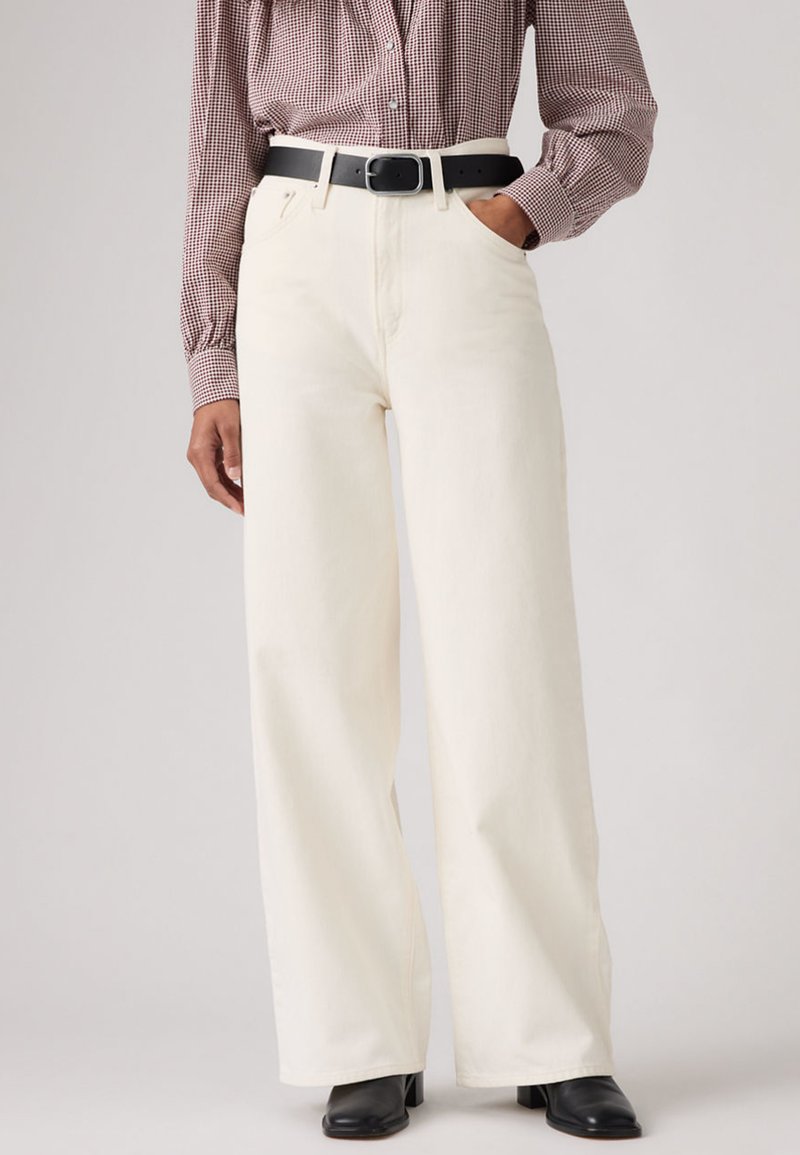 Γυναίκα που φοράει άσπρο παντελόνι με φαρδιές ίσιες γραμμές και ψηλή μέση, μαύρη ζώνη, μπορντό πουκάμισο με καρό σχέδιο και μαύρα μπότα με τακούνι, στέκεται με το ένα χέρι στην τσέπη.