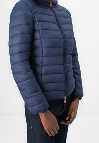 Person trägt eine navyblaue Steppjacke mit Reißverschluss und Fronttaschen sowie eine dunkle Jeans und steht vor einem schlichten weißen Hintergrund.