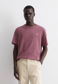 Camiseta de algodón burdeos con mangas cortas y cuello redondo, que presenta un pequeño logo blanco en el lado izquierdo del pecho. Combinada con pantalones beiges.