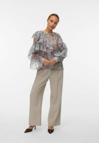 Blusa trasparente a più strati con motivo floreale in colori pastello, abbinata a pantaloni beige a gamba larga e scarpe con tacco marroni.