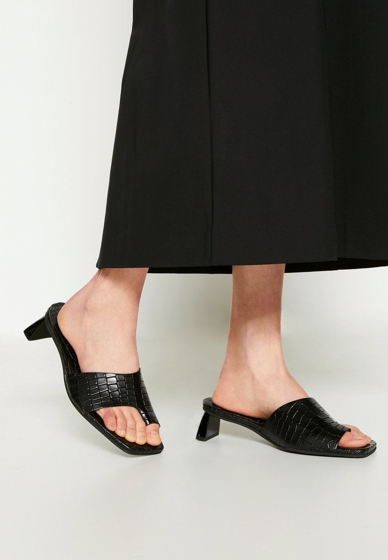 Personne portant un pantalon noir à jambes larges et des sandales mules noires ouvertes, à talon bas, avec un motif crocodile texturé.