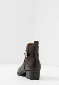 Bota de tobillo de cuero sintético marrón con tacón ancho, paneles laterales elásticos y una correa decorativa con un adorno circular con hebilla.