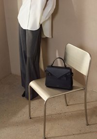 Borsa a mano in pelle nera con un design strutturato, caratterizzata da una patta frontale e doppi manici, poggiata su una sedia di metallo accanto a una persona in pantaloni grigi.