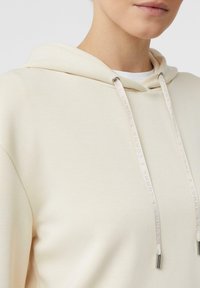 Felpa color crema con una texture morbida, dotata di un cordino con logo in rilievo e punte in metallo, e una vestibilità rilassata.