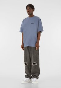 Tricou supradimensionat de culoare albastru deschis, cu o mică grafică neagră, asortat cu blugi gri cu rupturi și adidași albi cu accente gri.