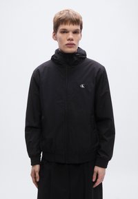 Μαύρο ελαφρύ μπουφάν με φερμουάρ μπροστά, κουκούλα, ελαστικά μανίκια και μέση. Διαθέτει μικρό λευκό λογότυπο στο αριστερό μέρος του στήθους.