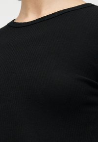 Κοντινό πλάνο ενός ατόμου που φοράει εφαρμοστό μαύρο πλεκτό ριμπ τοπ με στρογγυλή λαιμόκοψη σε ουδέτερο φόντο.