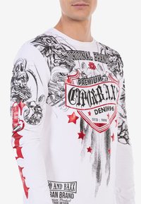 Langärmliges weißes T-Shirt mit schwarzen und roten Grafikdrucks, darunter Sterne, Totenköpfe und ein Wappenlogo mit dem Text "Premium Denim."