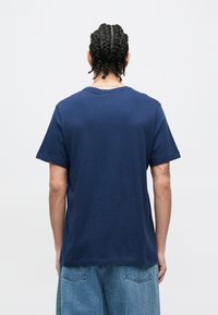 Μπλουζάκι από βαμβάκι σε ναυτικό μπλε χρώμα με κοντά μανίκια και στρογγυλή λαιμόκοψη. Η πίσω όψη δείχνει ομαλή υφή και ελάχιστες λεπτομέρειες ραφής.