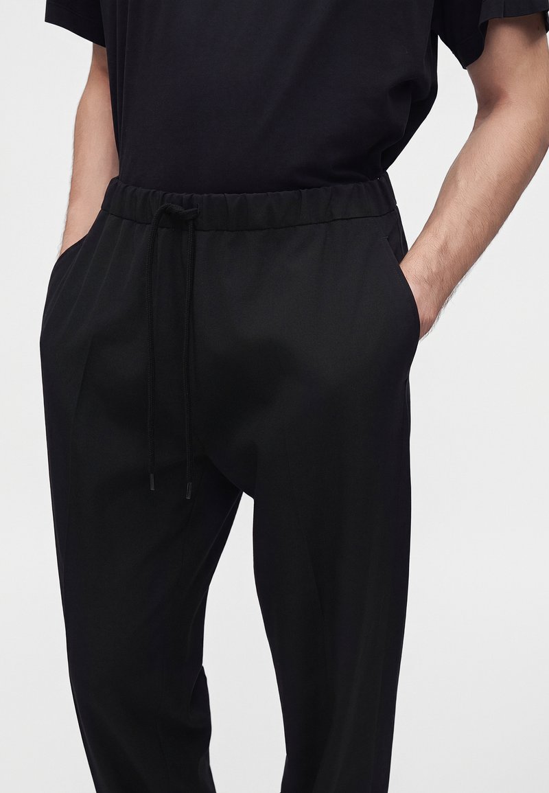 Personne portant un pantalon noir à cordon et une chemise noire à manches courtes, mains dans les poches, sur fond uni.