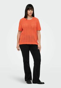 Gehaakte oranje trui met korte mouwen en een gestructureerd diamanten patroon; gecombineerd met zwarte flared jeans en zwarte schoenen.