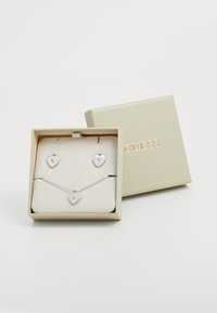 Silver heart-shaped necklace and earrings set inside a cream-colored box with gold "GUESS" logo. Box features smooth texture and clasp.
