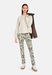 Helles graues, verkürztes Sweatshirt, Blumendruck-Hosen in Gelb- und Grüntönen und weiße Sneaker mit beigen Sohlen. Das Model hält eine braune Jacke.