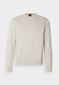 Pull col rond à manches longues couleur crème avec poignets et ourlet côtelés, avec une petite étiquette logo carrée sur la poitrine gauche.