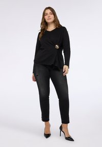 Top nero a maniche lunghe con scollo a V e dettaglio frontale attorcigliato, accentuato da una chiusura circolare dorata; abbinato a jeans skinny neri strappati e tacchi.