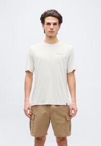 Νέος άνδρας που φοράει ανοιχτό μπεζ μπλουζάκι Patagonia και καμηλοπάρδαλο cargo σορτς, στάσιμος μπροστά σε απλό λευκό φόντο.