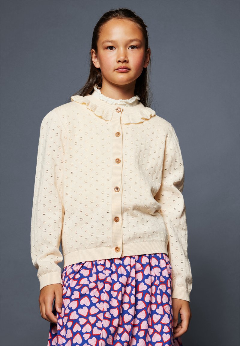 Cardigan a maglia color crema con colletto arricciato, caratterizzato da un motivo decorativo a occhiello e bottoni marroni. Abbinato a una gonna a cuore con motivo.