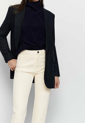 Kobieta w czarnej marynarce na granatowym, teksturowanym swetrze oraz kremowych, wysokim stanem spodniach, pozująca z jedną ręką w kieszeni.