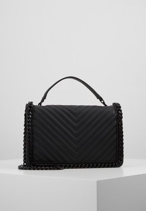 Sort quiltet skuldertaske med chevronmønster, top håndtag og kraftig kæderem, som hviler på en hvid sokkel mod en ensfarvet baggrund.
