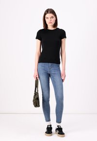 Tricou negru cu model canal, croi mulat, cu mâneci scurte; asociat cu blugi skinny albaștri deschiși; modelul ține o geantă verde și poartă adidași negri.