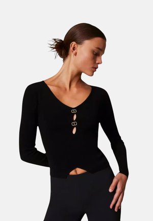 Mujer con el pelo castaño recogido lleva una camiseta negra de manga larga con dos aberturas decorativas con broches en el pecho y pantalones negros.