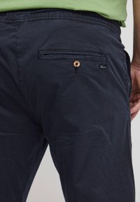 Navyblauwe broek met een achterzak voorzien van een houten knoop, subtiele textuur en een klein stoffen label. Materiaal lijkt lichtgewicht en casual.