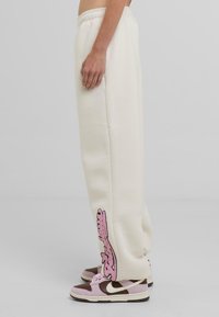 Crème kleurige sweatpants met een zijdesign in roze borduursel, met een geribbelde onderkant en een elastische tailleband. Gecombineerd met roze en bruine sneakers.
