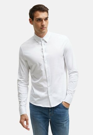 Mann trägt ein weißes Hemd mit langen Ärmeln und blaue Jeans, eine Hand in der Tasche, schaut leicht zur Seite.