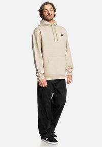 Beige hoodie met een voorzak en koorden, gecombineerd met losse zwarte broek en zwarte schoenen met witte zolen.