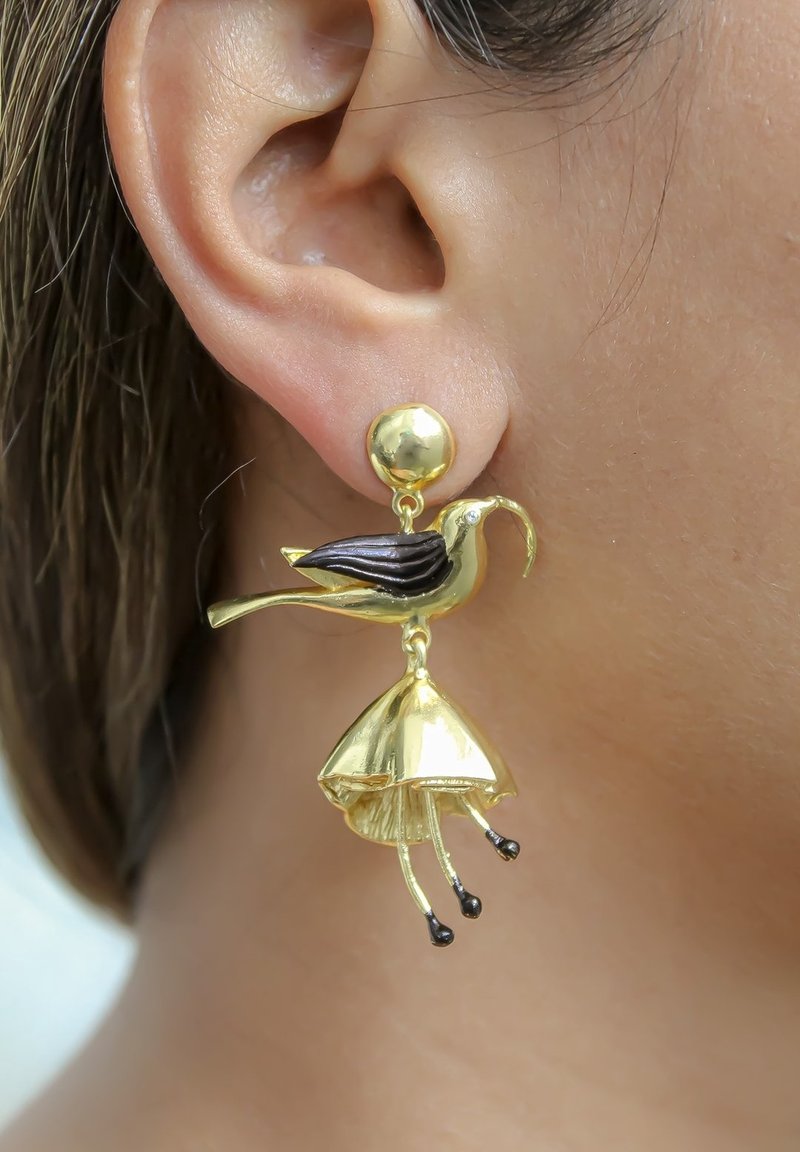 Boucle d'oreille dorée en forme d'oiseau au long bec tenant une fleur aux étamines noires, portée à l'oreille d'une personne.