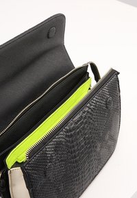 Černá kabelka se vzorem hadí kůže s texturovaným povrchem, v jasně zelené podšívce a zlatými zipovými detaily. Otevřená, aby ukázala vnitřní přihrádky.