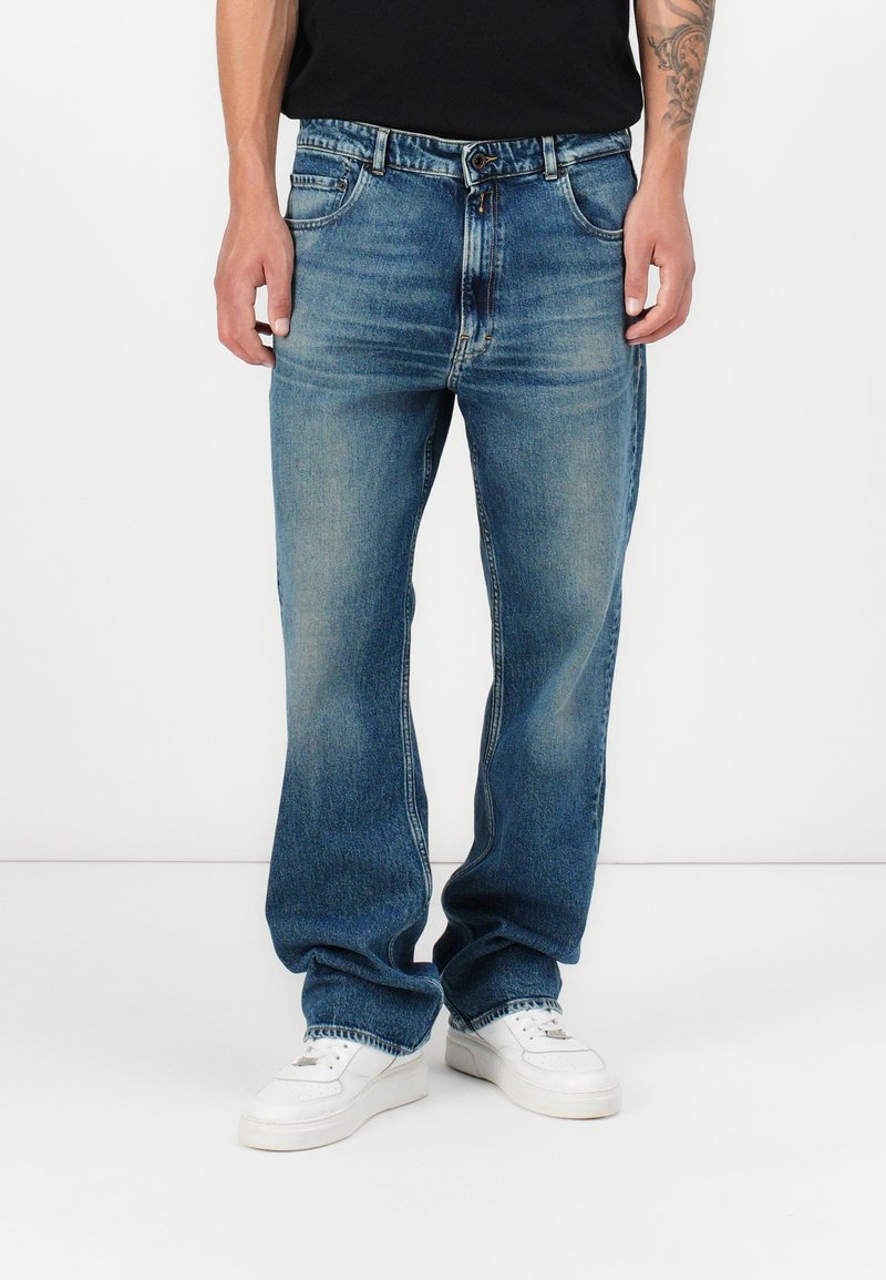 Blue denim jeans with a relaxed fit, featuring a faded finish, classic five-pocket design, and a button closure. Worn with white sneakers.