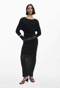 Zwarte geribbelde gebreide lange jurk met lange mouwen, voorzien van een zilverkleurige ronde riem. Draagt zwarte sandaalhakken met bandjes.