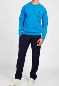 Sudadera azul con un bolsillo en el pecho con cremallera, combinada con pantalones azul marino. Zapatillas blancas completan el conjunto. Texturas de tela suaves y casuales.