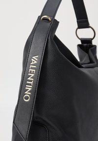 Črna usnjena torbica z teksturirano površino, z zlatim logotipom "VALENTINO" in ravnim naramnim trakom z metalnim obročem kot poudarkom.